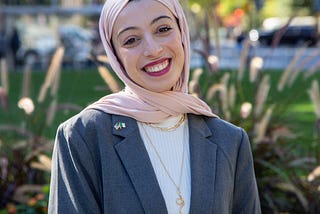 A woman in her 20s wearing a pink hijab is smiling at the camera, shown shoulders up. Behind her is a city park and buildings.