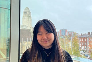 Lisa has long dark hair. The bottom half is dyed lighter and her fringe is swept to each side of her brow. She’s standing in front of a window, smiling, with a glimpse of campus and Parkinson tower peeking out from behind her. She’s wearing a black knit sweater and a gold pendant necklace.