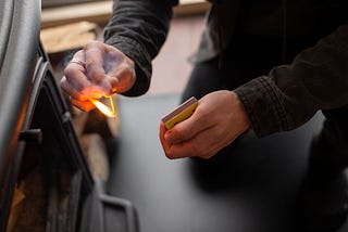 Photo of man’s hands holding a lit match, ready to light a wood stove. Courtesy of Freepik. https://www.freepik.com/free-photo/close-up-man-lighting-up-fire_22711891.htm#query=lighting%20a%20fire&position=8&from_view=search