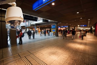 A large white dome surveillance camera is mounted indoors in the foreground, angled toward a busy transit concourse. In the background, slightly blurred, multiple people walk or stand beneath overhead signage and recessed lighting. The space has tiled floors, columns, storefronts, and illuminated signs, suggesting a train station or transportation hub.