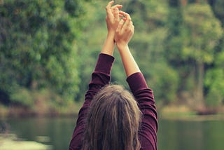 A long-haired woman, her back to us, stands before a lake, her arms up stretched.