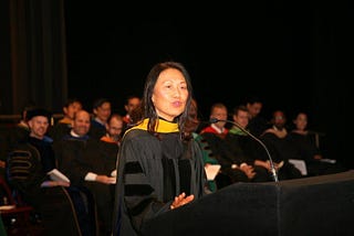 Woman dressed in academic regalia speaking at lectern with two seated rows of similarly dressed professors behind her.