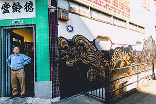 A photo of an Asian man standing in front of a store in Oakland Chinatown. There is a long golden and black dragon mural to the right of him. He is standing in front of a green wall, below a sign written in Chinese.