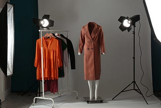 Professional clothing photography setup with studio lights, a mannequin displaying a brown coat, and a clothing rack holding colorful garments, all arranged in a well-lit studio with a grey backdrop.