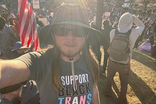The author is standing in front of a large crowd of people protesting. He’s wearing a hat and sunglasses. He has long brown hair and a beard. His t-shirt says “I support Black trans futures.” There’s an American flag behind him on the left and several protest signs. One sign says, “stop lying”. A person in the background is wearing a trans flag on the right. Another person in the background is waving a Mexican flag.