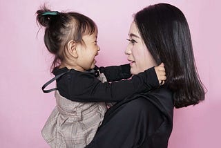 A woman with black hair wearing a black shirt holds a little girl with a ponytail against a pink background.