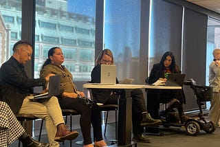 Estefania Cubillos Nova sitting on a panel with four other people. She is wearing black pants, a brown blazer and her hair is tied back. In front of her is a desk with a laptop.