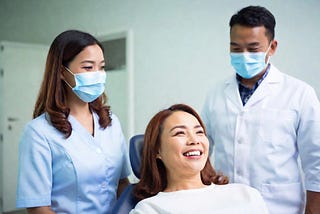 A female lie down on the wheelchair and two dentist stand side of patient one dentist is male and other female both wearing mask and dentist uniform