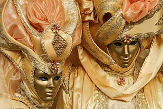 A man and woman in golden full-face masks and rich golden-and-peach-colored costumes.