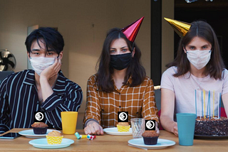 three people sit at a table with party hats and masks on. the table has cupcakes, NYU Local logos cover the candles.