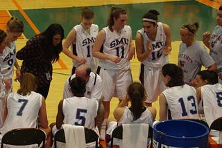 A photo of women on a basketball court, huddled around their couch during a timeout.