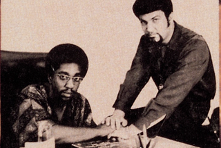 Two black males. The picture is at a desk. Barrett is sitting in the chair at the desk and Whitfield is sitting on the desk to his left. The men have their hands one on top of the others hand interlocked. Barrett is in glasses and a plaid shirt. Whitfield is in a solid shirt with no glasses.