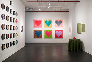 An art gallery space is pictured that contains 26 colorful clocks, multiple colorful framed prints of hearts and squares, and a fabric installation with a tablecloth and matching stretched canvases.