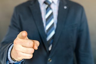 A person with coat and tie pointing his index finger towards the camera