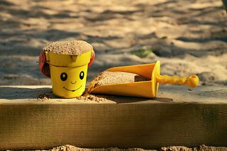 A sand box with a yellow bucket, that shows a smiling face, and a shovel next to it.