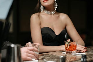 A close-up shot of an elegant woman sitting at a bar with an orange cocktail in front of her. She wears a strapless black dress, diamond drop earrings, and a choker necklace. We only see her from her lips to her arms resting on the bar. To the left is a man’s hand holding a glass.