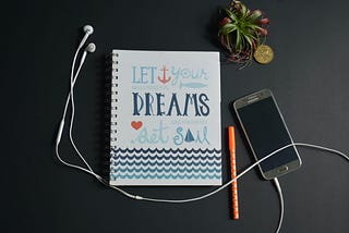 A mobile phone with attached headphones accompanied by notebook with title that reads “Let Your Dreams Set Sail.”
