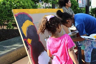 two young brown girls painting a woman on a canvas