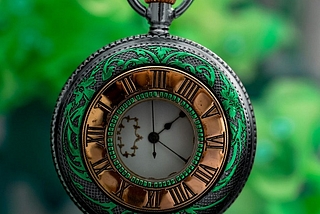 A small antique clock for a chain suspended over a green leafy background. The face is white, and the roman numerals are located on a gold band around the face. The frame is a dark gray silver with a decorative green detail.