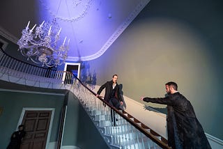 Two men in black suits stand at the top of a dramatically-lit staicase. One man grasps the railing with both hands as he backs up the stairs. The other man approaches him from a few steps down, one arm outstretched.