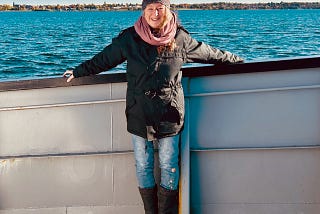A woman stands on the deck of a boat. Water stretches behind her. Her arms are resting on the siderails.