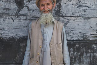 An older man with a white beard, smiling and standing against a grey background.