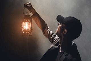 A man in a black ballcap holding a lit lantern up towards the darkness.