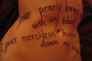 A woman’s belly with words written in ink pen on her skin, “your pearly horns, with my blood, your merciless hands, down my walls.”