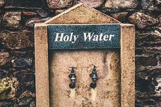 Colour image of two ordinary taps/faucets located in an outside wall and above them is a sign with the words ‘Holy Water’.