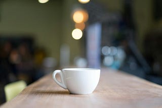 A white coffee cup sitting on a wooden table in a cozy café, with warm blurred lights in the background.