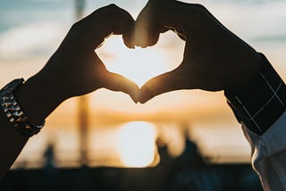 Two hands forming a heart. The sun is setting in the background.