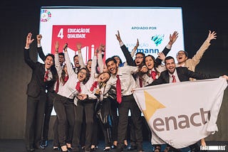 Grupo de universitários com roupa social e gravata vermelha comemorando sobre um palco