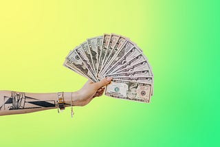 An outstretched arm with a tattoo on it is holding several 5, 10, and 20 dollar bills in a fan formation.