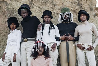 Six Black people standing outside of a rock formation wearing 80s style fashion