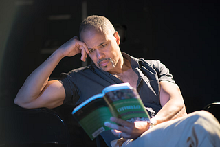 A Black man with short hair and a moustache in a short-sleeved gray shirt sits and reads Othello.