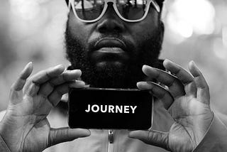 A black and white portrait of a cool man in a Nike cap and mirrored sunglasses holding a phone with the word ‘journey’ on the screen