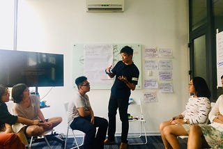 Liang-Yi Chang standing in front of a wall full of handwritten posters, gesturing with his hands as he speaks to five people sitting in plastic folding chairs.