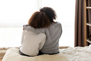 Mother and daughter sit on the bed, the girl’s head leaned against her mom