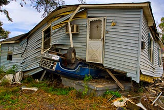 An unusual scene, with a damaged and twisted house resting on top of a blue vehicle that is upside down.