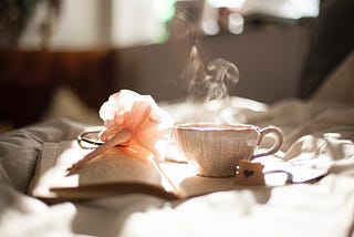 ceramic teacup with steaming coming out of it on open notebook beside pink flower pen on bedside