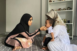 In the image, two women and a young child are seated on a patterned carpet indoors. One woman, dressed in a black and pink outfit with a black hijab and glasses, is gently touching the face of the child, possibly in a comforting or affectionate gesture. The child, who appears to be a toddler, is sitting on the lap of the second woman, who is wearing a white outfit and a light-colored patterned hijab. Behind them is a white shelf containing various decorative plates and items.