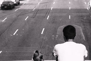 A picture of a father and young daughter, sitting on bridge, overlooking urban traffic.