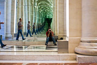 An “infinite mirror” image of a man walking into a columned hallway. Another man, seated and holding a camera to his eye, faces the viewer,