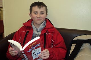 A child around ten years old reading “Diary Of A Wimpy Kid.” He is sitting on a black sofa with a cream-colored wall behind him. He’s wearing a grey hoodie and red jacket. He has brown hair.