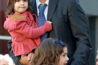 Adam Sandler is in a suit and tie, holding one of his daughters who has short brown hair and is wearing a pink top, his other daughter is standing in front of him she has longer curly hair and is in a blue and white striped top.