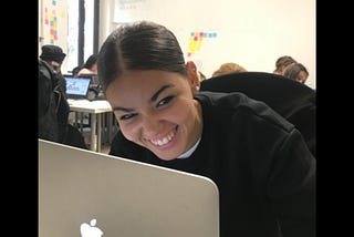 Fazia Bouheaoura, a web developer smiling graciously, sitting in of front of her laptop working in a room filled with women.