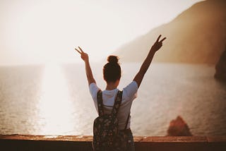 woman with backpack standing near sunny seashore with peace signs in the air