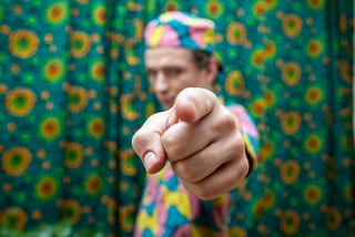 A man standing in front of a colorful curtain backdrop. He’s wearing an outfit with a colorful pattern and is pointing directly at the camera.