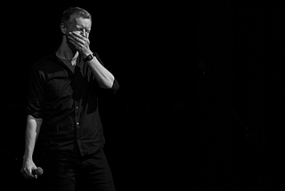 an improviser standing on stage with his eyes closed, holding a microphone in one hand and covering his mouth with the other