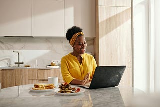 A Black woman in a yellow shirt and polkadot hair scarf smiling at a computer at her kitchen table, with breakfast next to her